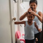 Une femme coiffe une petite fille souriante devant un petit pot rose dans une salle de bain